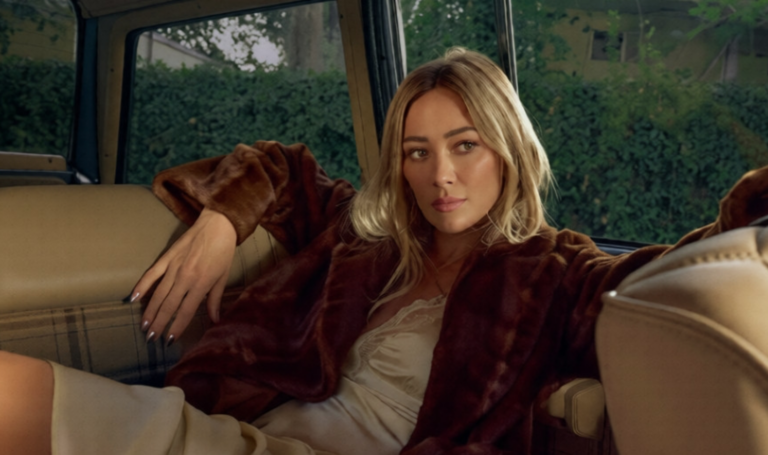 Hilary Duff siede sul sedile posteriore di un'auto indossando una giacca di pelliccia marrone sopra un abito color crema
