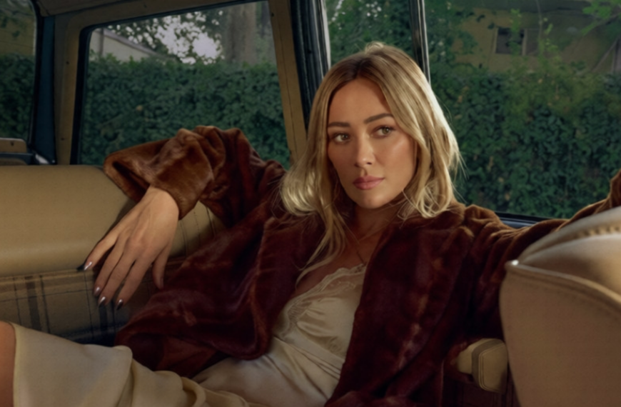 Hilary Duff siede sul sedile posteriore di un'auto indossando una giacca di pelliccia marrone sopra un abito color crema