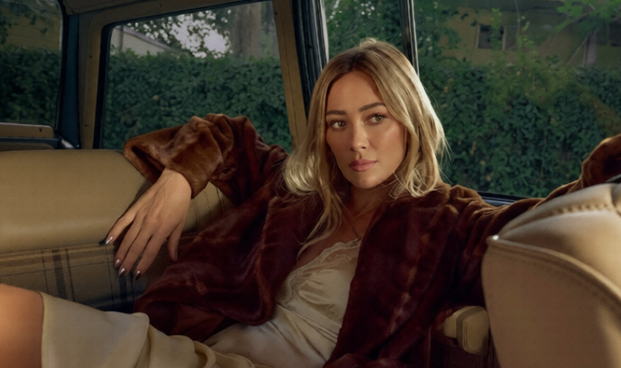 Hilary Duff siede sul sedile posteriore di un'auto indossando una giacca di pelliccia marrone sopra un abito color crema