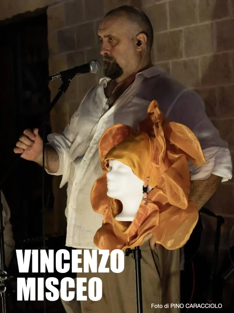 Vincenzo Misceo canta al microfono tenendo in mano una testa di manichino con un copricapo arancione ispirato ai Genesis