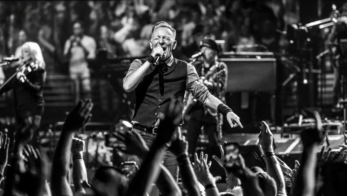 Bruce Springsteen sul palco in bianco e nero mentre canta al microfono davanti a un pubblico con mani alzate e musicisti