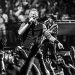 Bruce Springsteen sul palco in bianco e nero mentre canta al microfono davanti a un pubblico con mani alzate e musicisti
