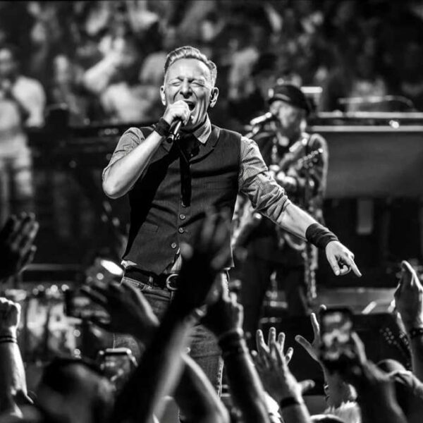 Bruce Springsteen sul palco in bianco e nero mentre canta al microfono davanti a un pubblico con mani alzate e musicisti