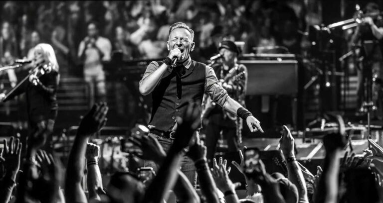 Bruce Springsteen sul palco in bianco e nero mentre canta al microfono davanti a un pubblico con mani alzate e musicisti