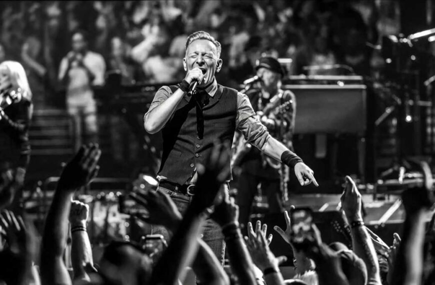 Bruce Springsteen sul palco in bianco e nero mentre canta al microfono davanti a un pubblico con mani alzate e musicisti