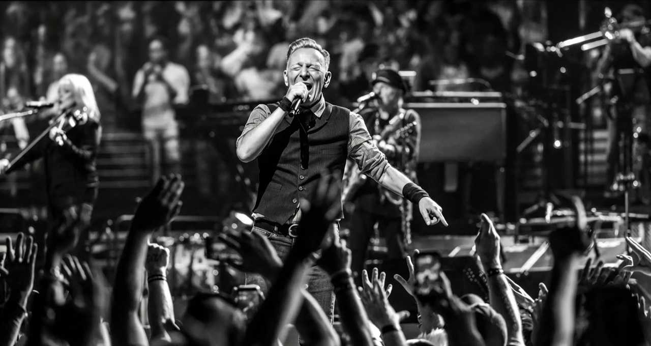 Bruce Springsteen sul palco in bianco e nero mentre canta al microfono davanti a un pubblico con mani alzate e musicisti