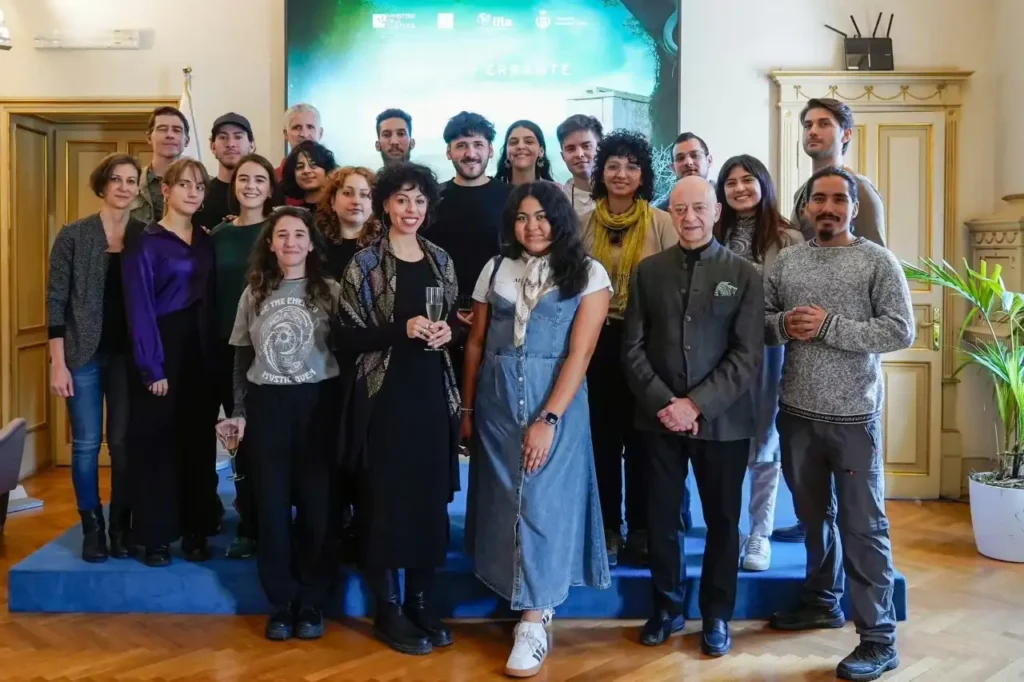 Gruppo di persone in piedi su un palco blu in una sala storica, alcune sorridenti, una donna tiene un bicchiere e sullo