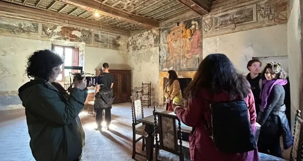 Persone con giacche e zaini che visitano una sala storica con affreschi, soffitto a cassettoni e una fotografa a sinistra