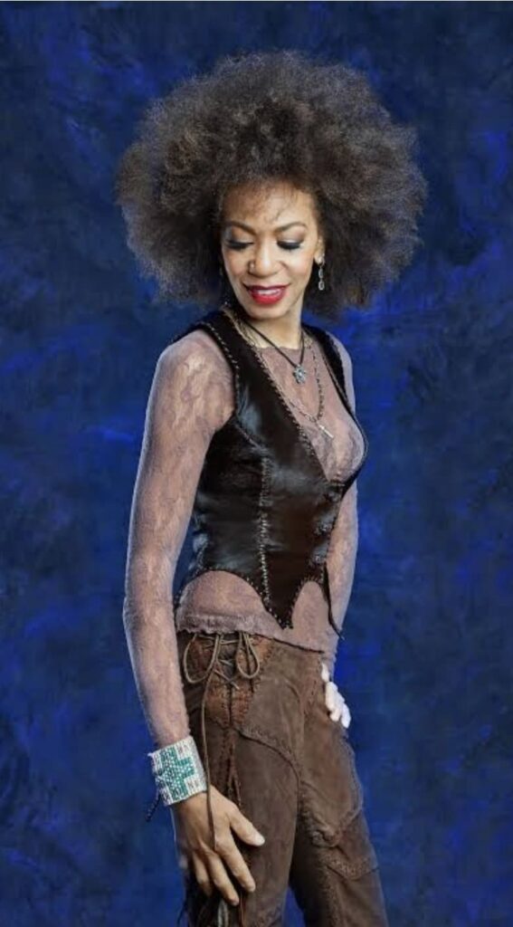 Cindy Blackman Santana con capelli afro, gilet in pelle e pantaloni marroni posa davanti a uno sfondo blu per un'intervista