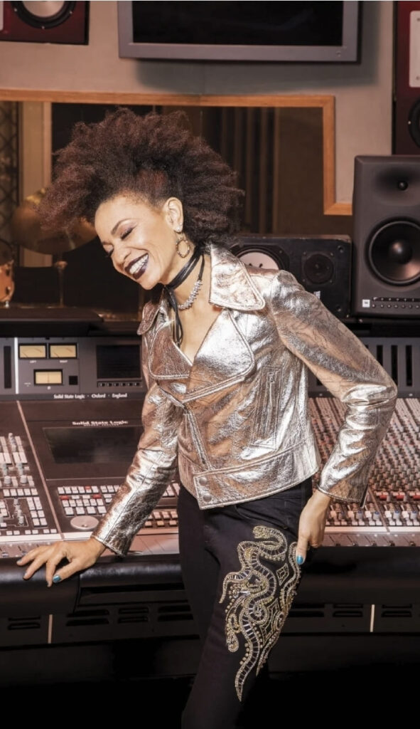 Cindy Blackman Santana sorride appoggiata a una console di registrazione in studio, indossando una giacca argento e