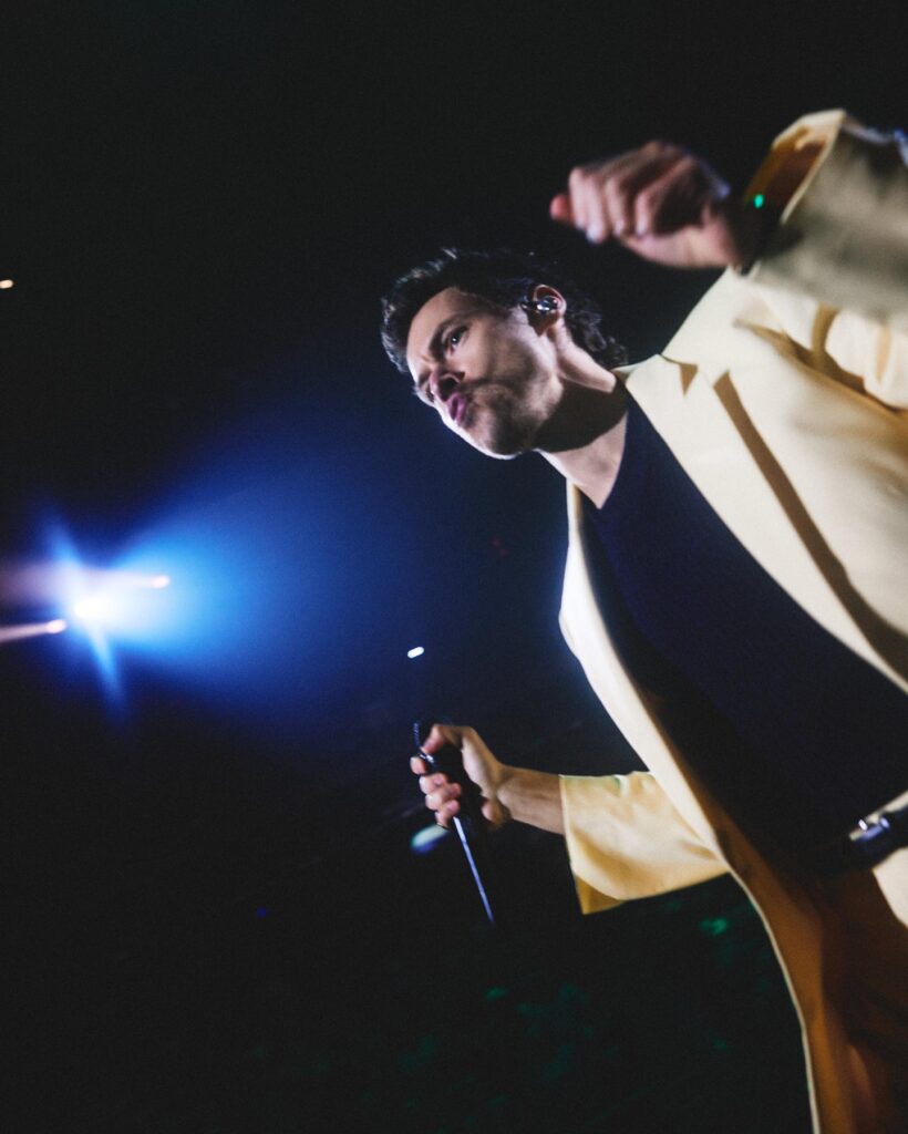 Harry Styles canta sul palco con una giacca gialla e un microfono in mano durante un'esibizione dal vivo