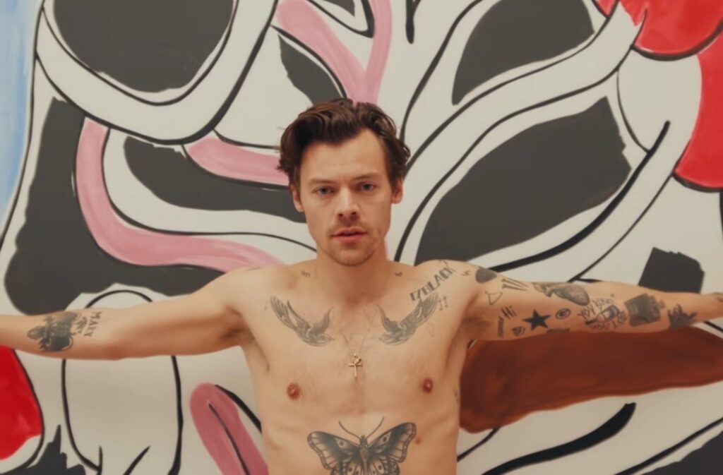 Harry Styles a torso nudo con tatuaggi in primo piano davanti a un grande dipinto astratto con forme nere, bianche e rosse