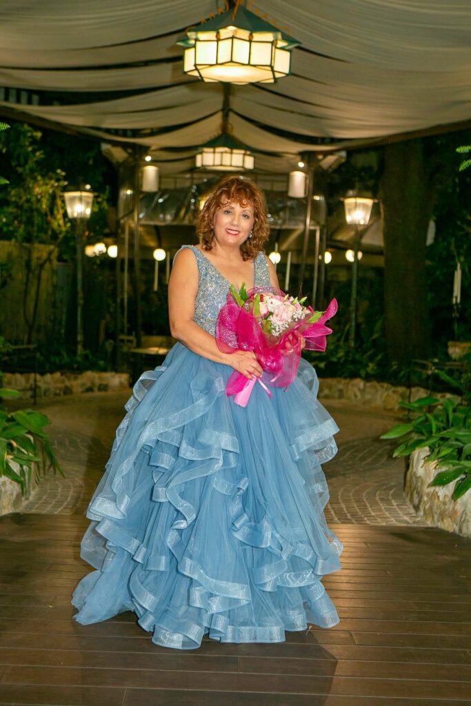 Maria Pia Cappello sorride indossando un elegante abito azzurro con gonna a balze mentre tiene un bouquet per il suo