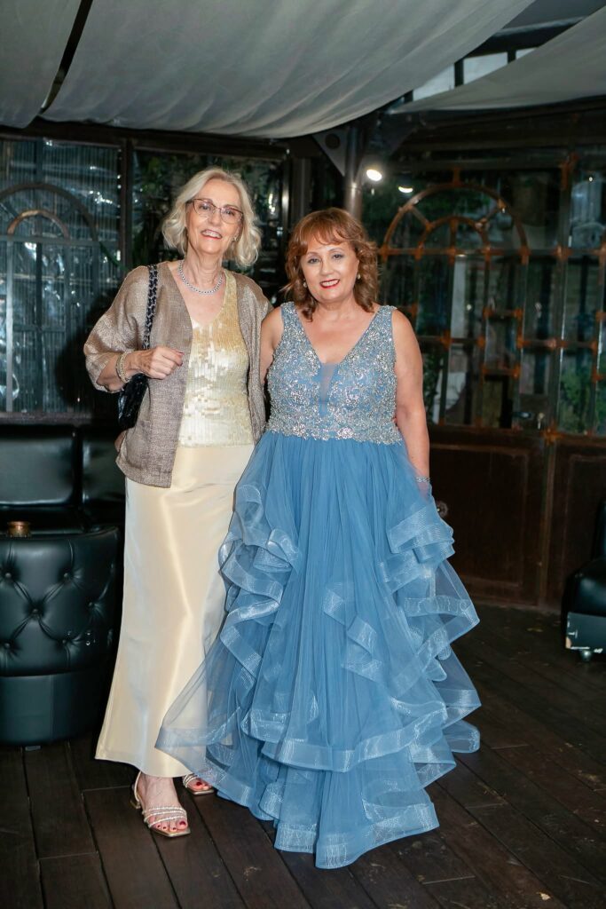 Donna sorridente in abito lungo azzurro con corpetto ricamato e balze tulle, accanto a donna con gonna crema e giacca
