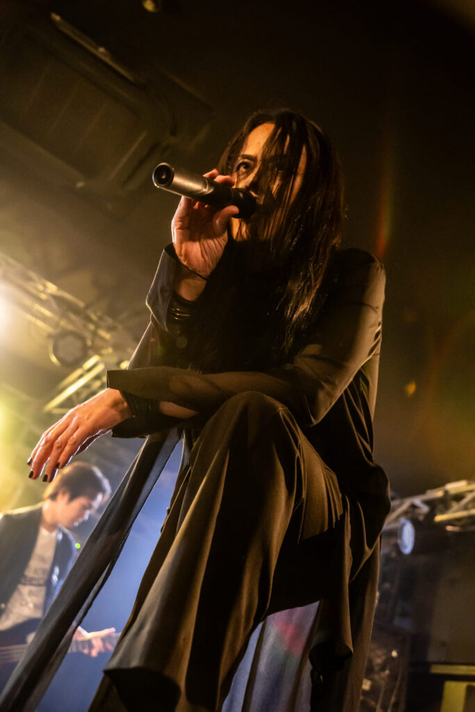 Koichi dei Neues in primo piano con microfono, capelli neri lunghi, abito scuro, posa accovacciata sul palco illuminato