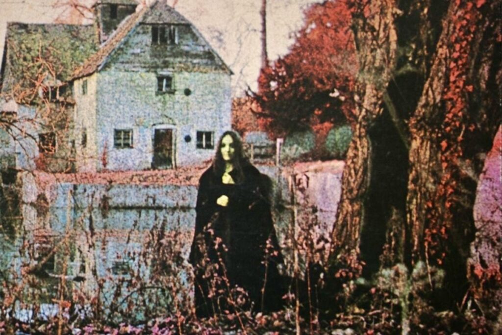 Donna in mantello nero in primo piano davanti a un casale bianco e uno stagno, alberi e foglie autunnali rosse e marroni