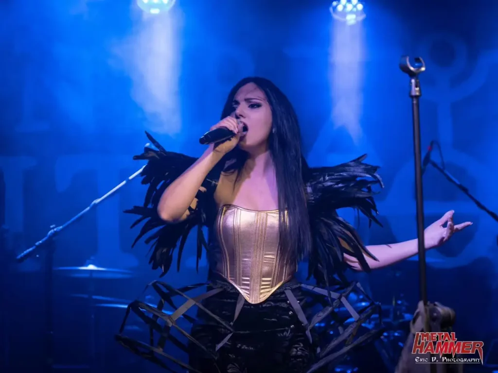 Irene Petitto sul palco con microfono, corsetto dorato, spalline piumate nere e luci blu durante un concerto