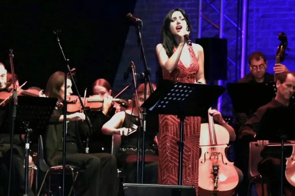 Irene Petitto canta al microfono indossando un abito rosso lungo, accompagnata da un'orchestra con violini e violoncello