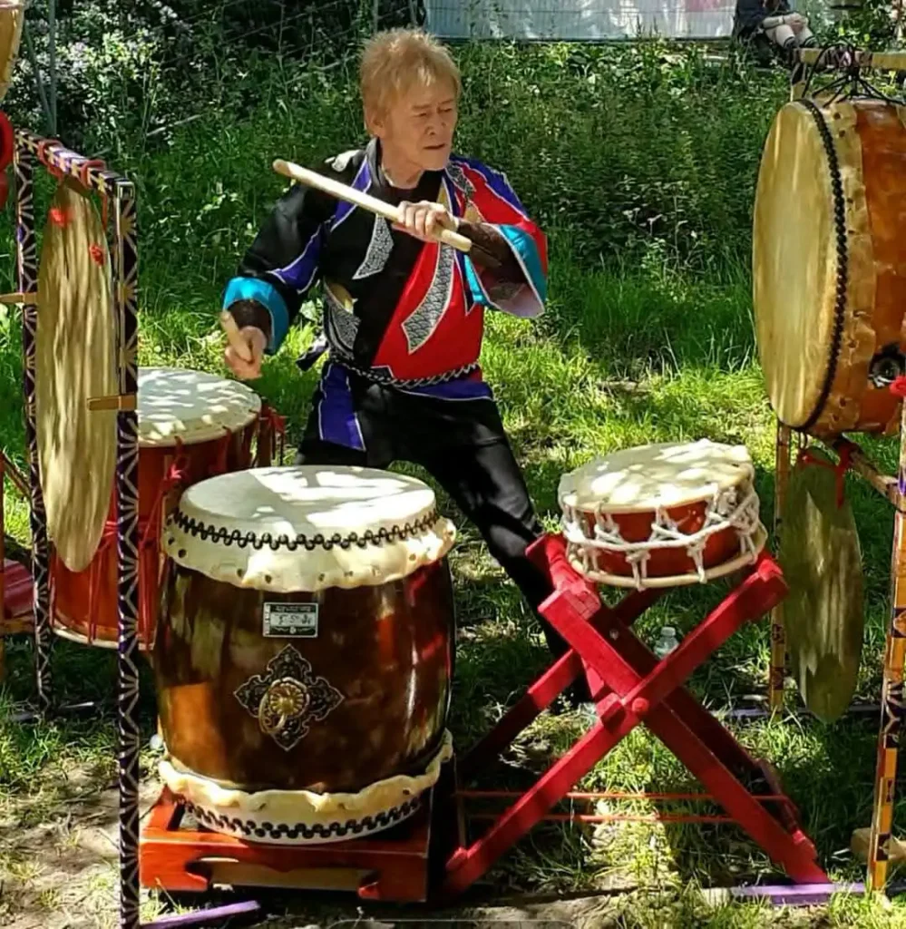 Joji Hirota suona un grande tamburo taiko in un giardino, indossando un tradizionale abito colorato rosso, blu e nero