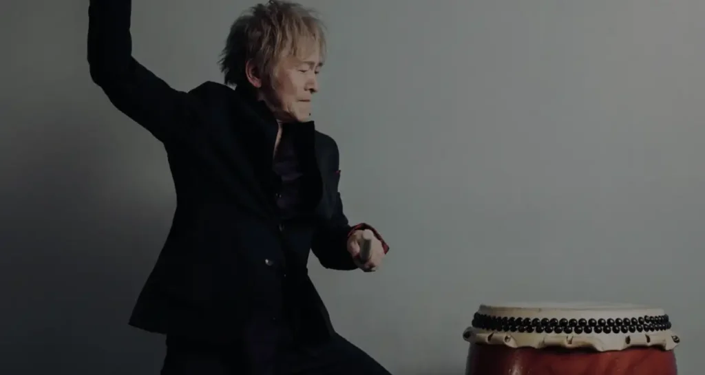 Joji Hirota indossa un completo nero mentre solleva una bacchetta per suonare un tamburo tradizionale giapponese taiko