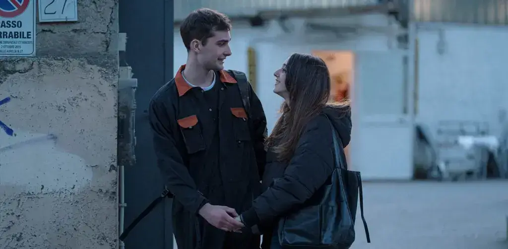 Un giovane uomo in tuta da lavoro e una ragazza si tengono per mano guardandosi intensamente in una scena del film La città