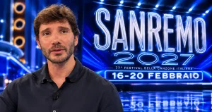Stefano De Martino in primo piano davanti al logo luminoso del Festival di Sanremo 2027 con le date 16-20 febbraio