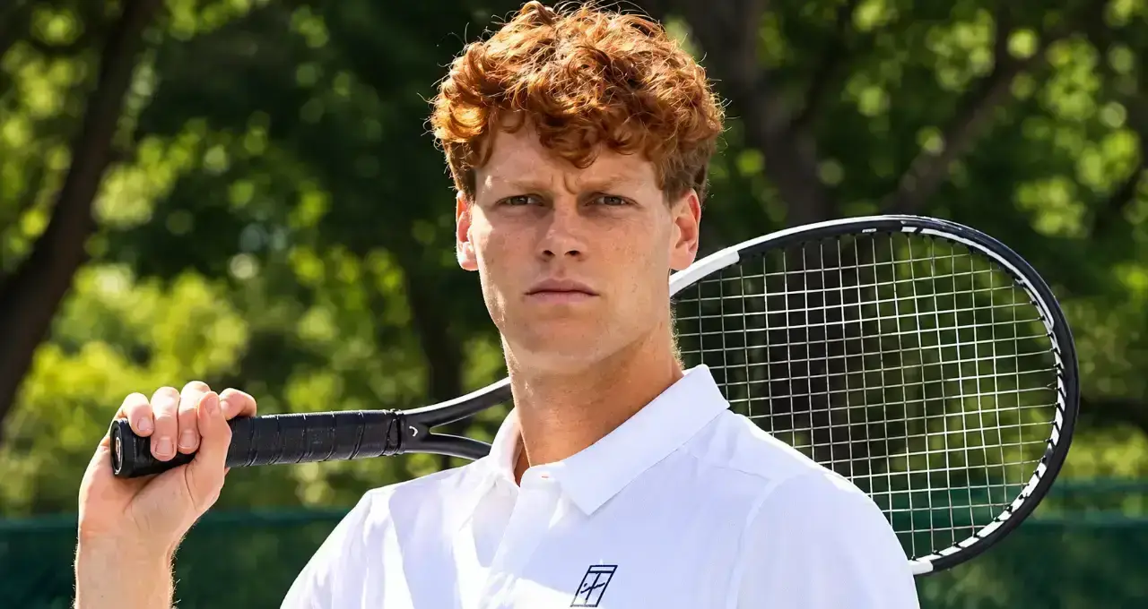 Jannik Sinner in polo bianca con racchetta appoggiata sulla spalla, sguardo serio e sfondo alberato del campo da tennis