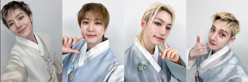 Quattro membri dei Stray Kids in hanbok chiari su sfondo neutro, sorridenti e in pose con mano al volto e pollice alzato