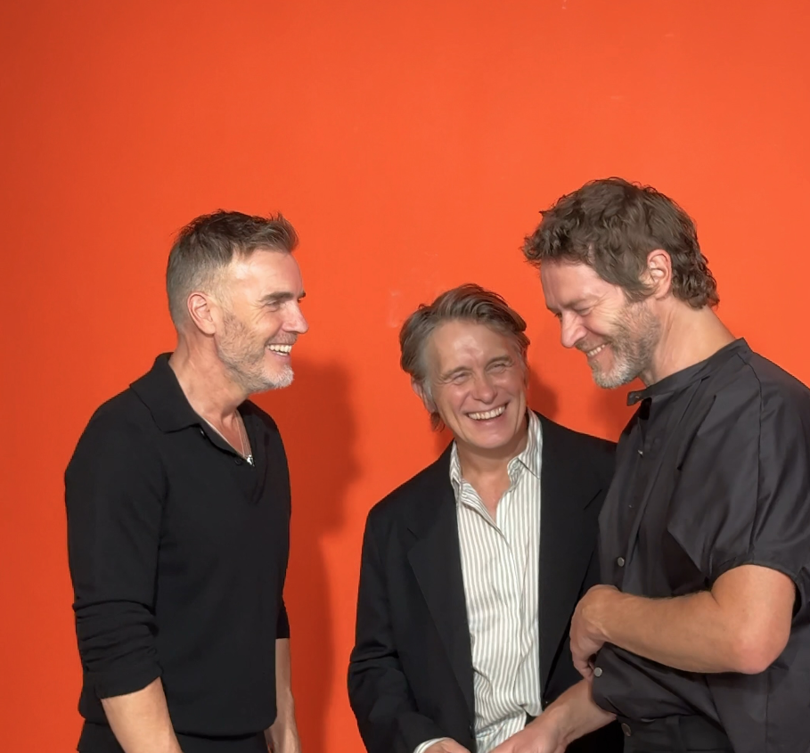 Gary Barlow, Mark Owen e Howard Donald dei Take That ridono insieme davanti a uno sfondo arancione per il singolo Higher