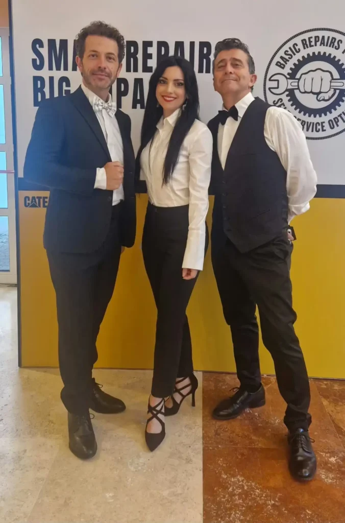 Irene Petitto in camicia bianca e pantaloni neri sorridente al centro tra due uomini in abito scuro davanti a un pannello
