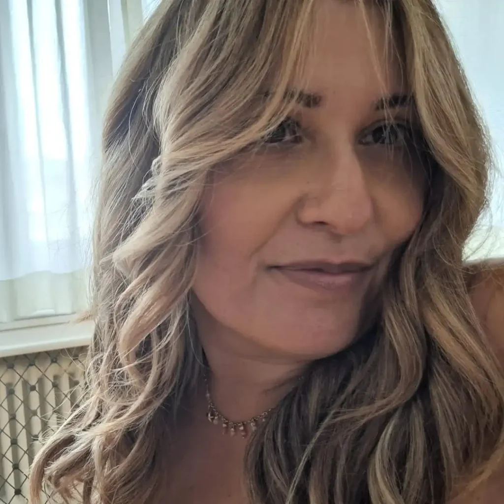 Donna sorridente in primo piano con capelli biondo cenere mossi, collana sottile e sfondo con tenda chiara e termosifone