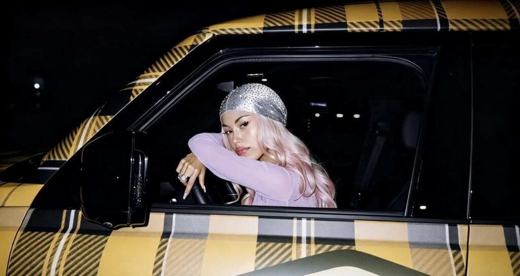 La rapper ANNA con capelli rosa e durag scintillante è seduta al posto di guida di un'auto con carrozzeria a quadri gialli