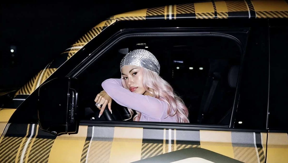 La rapper ANNA con capelli rosa e durag scintillante è seduta al posto di guida di un'auto con carrozzeria a quadri gialli