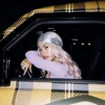La rapper ANNA con capelli rosa e durag scintillante è seduta al posto di guida di un'auto con carrozzeria a quadri gialli