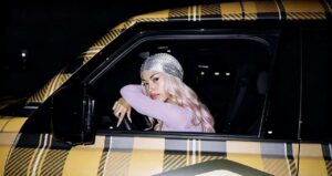 La rapper ANNA con capelli rosa e durag scintillante è seduta al posto di guida di un'auto con carrozzeria a quadri gialli