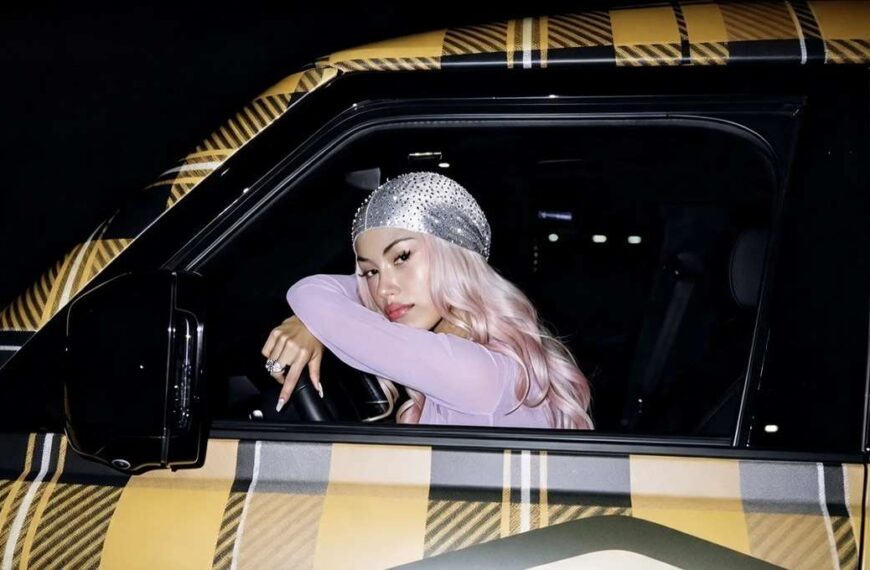 La rapper ANNA con capelli rosa e durag scintillante è seduta al posto di guida di un'auto con carrozzeria a quadri gialli