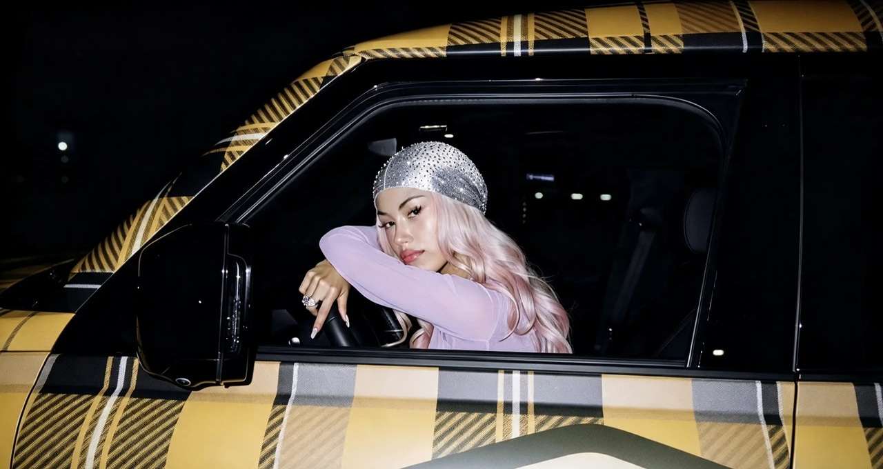 La rapper ANNA con capelli rosa e durag scintillante è seduta al posto di guida di un'auto con carrozzeria a quadri gialli