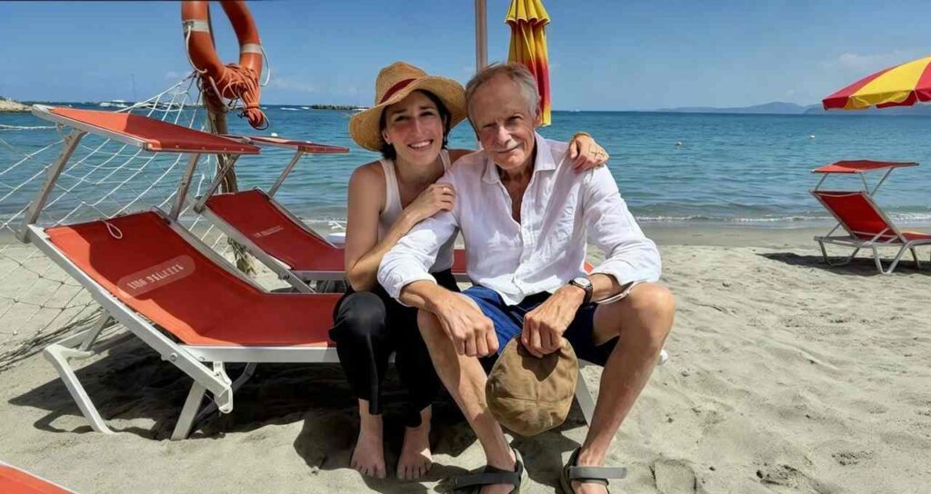 Donna con cappello di paglia e uomo anziano seduti sulla sabbia davanti a lettini arancioni e ombrelloni, mare calmo sullo