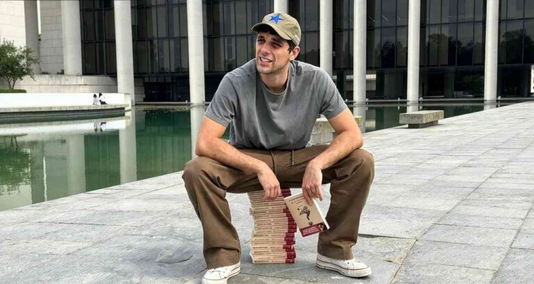 Il cantante Bresh seduto su una pila di libri mentre tiene in mano il suo romanzo Nessuno ci insegna a cadere