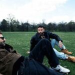 Cesare Cremonini e Salmo seduti sull'erba in un parco per la collaborazione Amateur, vestiti in stile casual e moderno