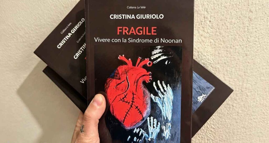 Una mano tiene in primo piano il libro Fragile di Cristina Giuriolo, con un cuore rosso disegnato in copertina