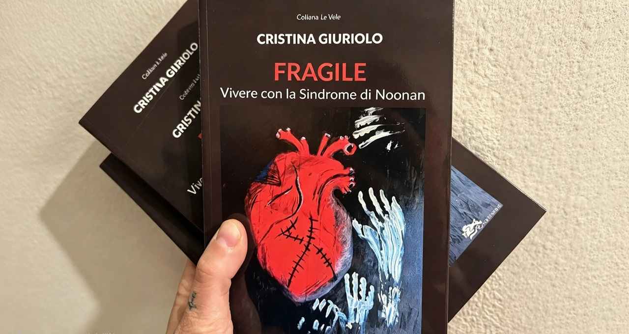 Una mano tiene in primo piano il libro Fragile di Cristina Giuriolo, con un cuore rosso disegnato in copertina