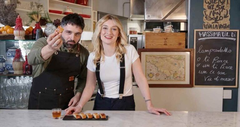 Ruben Bondì e una donna in cucina presentano dei piccoli stuzzichini su un vassoio davanti a una lavagna con scritte in