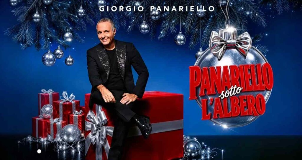 Giorgio Panariello seduto su un grande pacco regalo rosso, abito nero brillante, sfondo blu con decorazioni natalizie