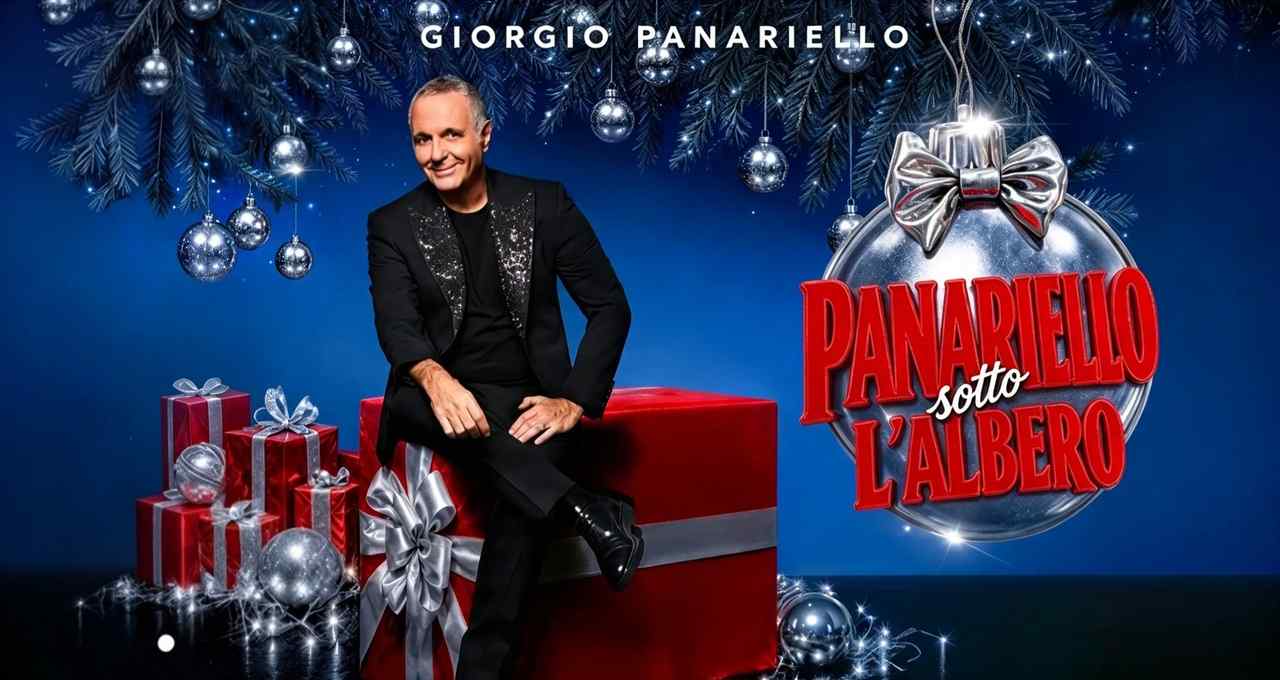 Giorgio Panariello seduto su un grande pacco regalo rosso, abito nero brillante, sfondo blu con decorazioni natalizie