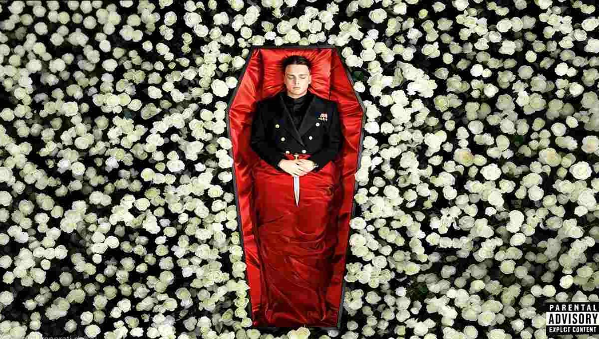 Uomo in uniforme scuro sdraiato in una bara rivestita di raso rosso, con un pugnale poggiato sulle mani, circondato da