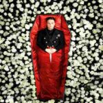 Uomo in uniforme scuro sdraiato in una bara rivestita di raso rosso, con un pugnale poggiato sulle mani, circondato da