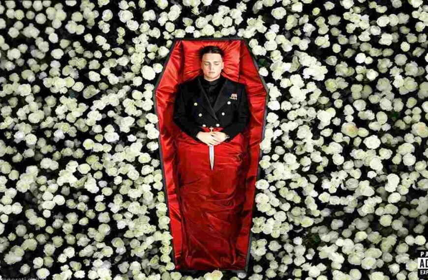 Uomo in uniforme scuro sdraiato in una bara rivestita di raso rosso, con un pugnale poggiato sulle mani, circondato da