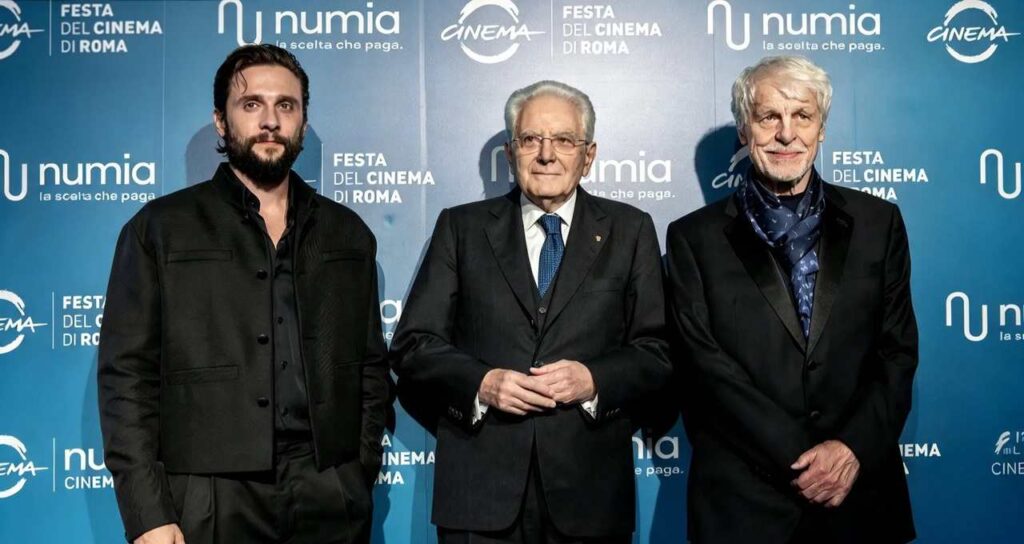 Tre uomini in abito scuro davanti a un backdrop blu con loghi "numia" e "Festa del Cinema di Roma", il centrale con cravatta