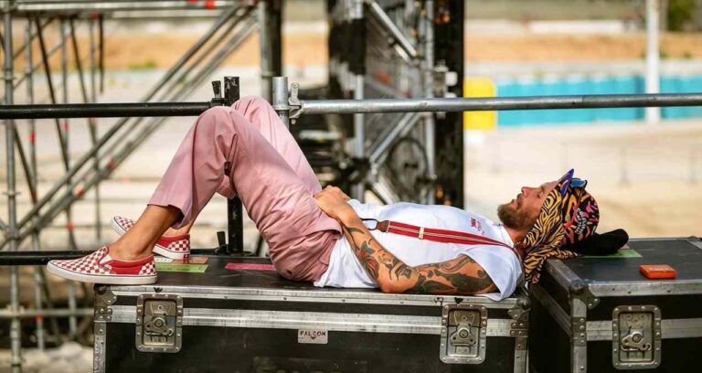 Jovanotti riposa sdraiato su un flight case nero con pantaloni rosa, t-shirt bianca, bretelle e scarpe a scacchi rosse